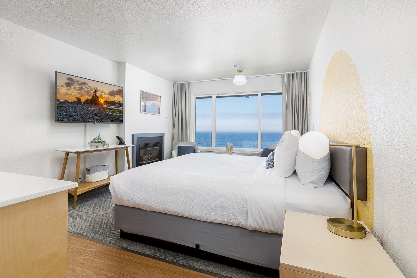 A modern hotel bedroom with a large bed, ocean view through windows, a wall TV, fireplace, and neutral decor.