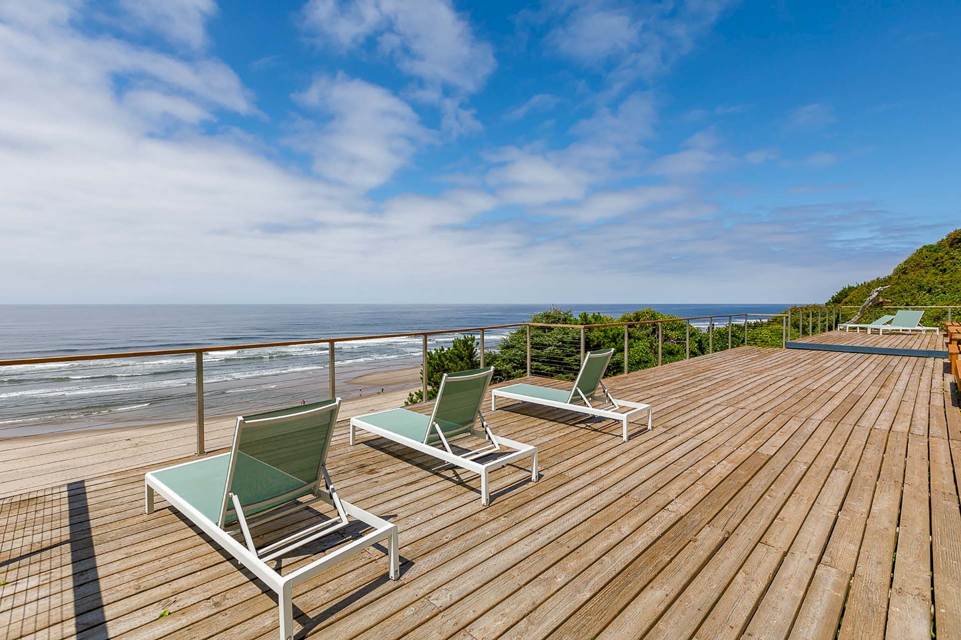 A seaside wooden deck with green lounge chairs and railings, overlooking a sandy beach, rolling waves, and a blue sky with clouds.