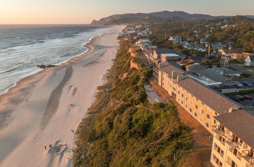 Aerial view of a coastal town at sunset, with a sandy beach, ocean waves, and hillside homes lining the shoreline.