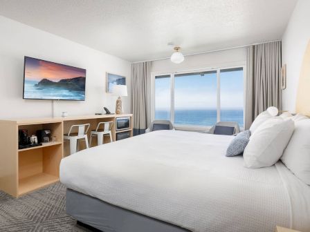 A bright modern hotel room with a large bed, TV, and ocean view through big windows.