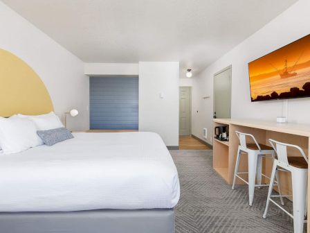 Modern hotel room: white bed, small bar with stools, and a sunset ship picture on the wall.
