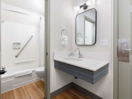 A bright modern bathroom with a white vanity and sink, round mirror, towels, and a bathtub in view.