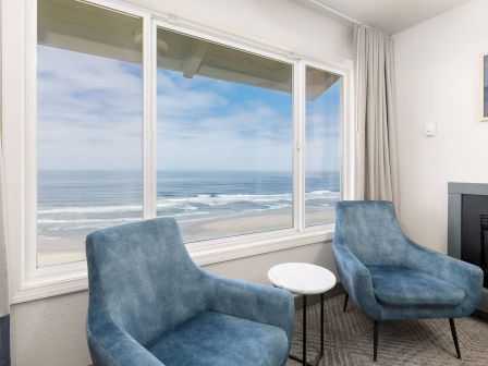 A bright seaside living room with two blue armchairs, a small white table, and wide windows showing ocean waves.
