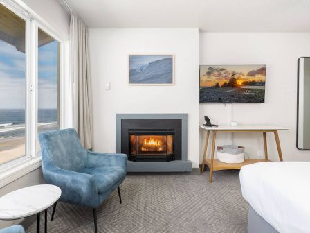 A bright coastal living room with ocean view, blue armchair, lit fireplace, wall art, desk, and a standing mirror.