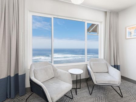 A modern room with two beige lounge chairs facing a large window showing an ocean and waves under a blue sky.
