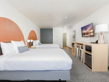 A modern hotel room with twin beds, a desk, TV, lamps, and neutral decor.