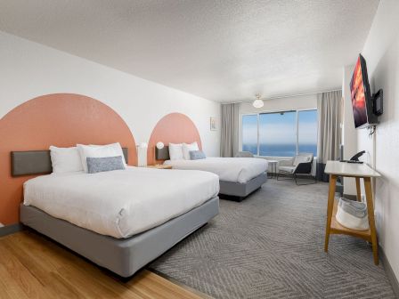 Modern hotel room with two beds, ocean view window, and a wall-mounted TV.