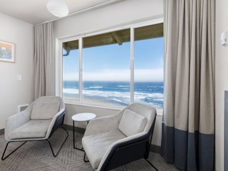 Bright lounge by ocean-view windows with two gray chairs, a small white table, and floor-to-ceiling curtains beside a fireplace.