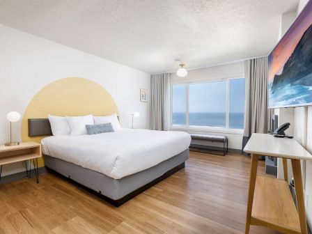 A modern bedroom with a large bed, bedside tables, curtains, ocean-view window, and a TV with desk by the wall.