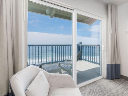 A bright living room with beige curtains and sofa, opening onto a balcony with ocean views and white railings.