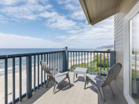 A seaside balcony with two white chairs and a table, overlooking the ocean and sandy beach under a partly cloudy sky.