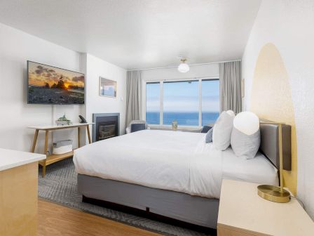 A bright hotel bedroom with a king bed, gray curtains, a fireplace, and a view of the ocean through large windows.