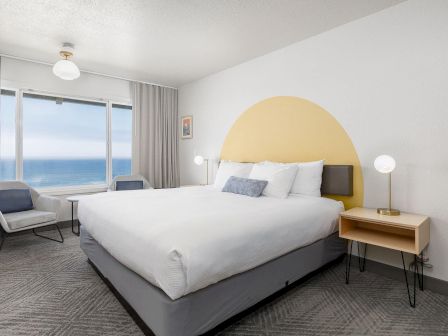A modern bedroom with a large bed, bedside tables, gray curtains, and a bright yellow arched accent wall overlooking the ocean.