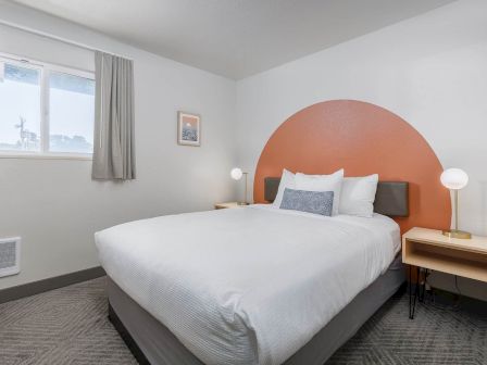 A tidy hotel bedroom with a white bed, orange arched headboard, two lamps, window, and framed wall art.