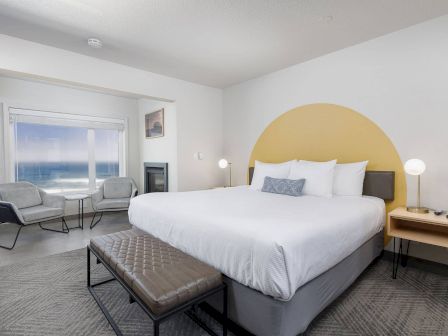 A modern hotel bedroom with a large bed, yellow arched headboard, bedside lamps, and a seating area by big windows.