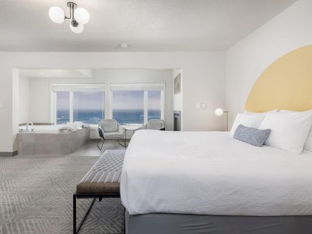 A bright modern hotel bedroom with a large bed, ocean-view windows, and a cozy seating area, lit by a ceiling chandelier.