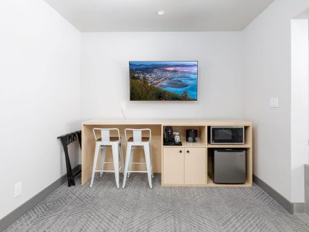 A clean modern kitchenette with two white bar stools, a cabinet bar area, a microwave/coffee setup, and a framed seaside photo.