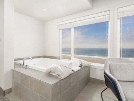 A bright seaside bathroom with a large jetted tub, sink, and a chair by big windows showing the ocean.