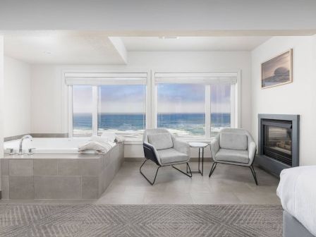 A bright seaside lounge with a soaking tub, two gray chairs, a small table, and an ocean view by a fireplace.