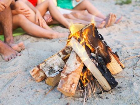A beach bonfire with burning logs while friends lounge nearby on the sand.
