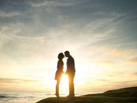 Silhouetted couple stands on a cliff by the ocean at sunset, sharing a quiet moment under colorful clouds.