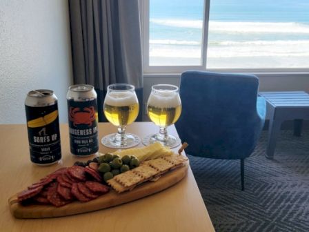 A table by the window holds four beer cans and two filled beer glasses, plus sliced meats, crackers, fruit, and olives, overlooking the sea.