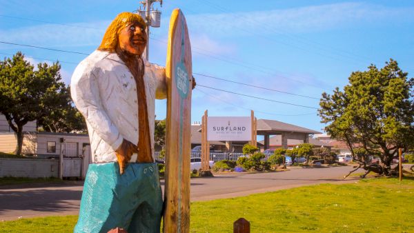 A large surfer statue by a roadside, with the Surfland Hotel by the Sea sign in the background.