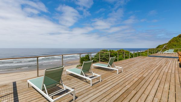 Oceanfront wooden deck with lounge chairs, railing, and a view of the blue sea under a cloudy sky.