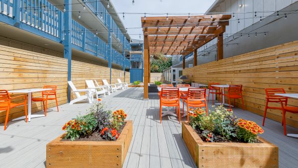 A sunny outdoor courtyard with string lights, wooden planters full of flowers, and bright orange-and-white chairs.