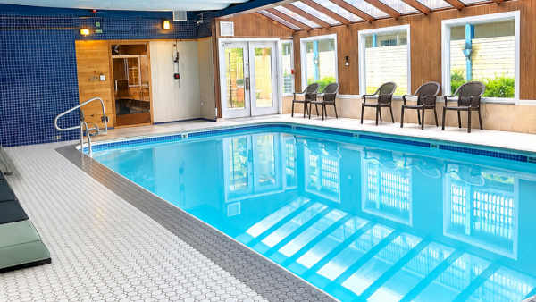 An indoor swimming pool with blue tiles, mosaic floor, wooden walls, and lounge chairs under a white ceiling canopy.
