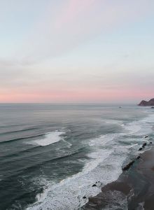 A quiet ocean shoreline at sunset with gentle waves, a sandy beach, rocky cliffs, and a distant headland.
