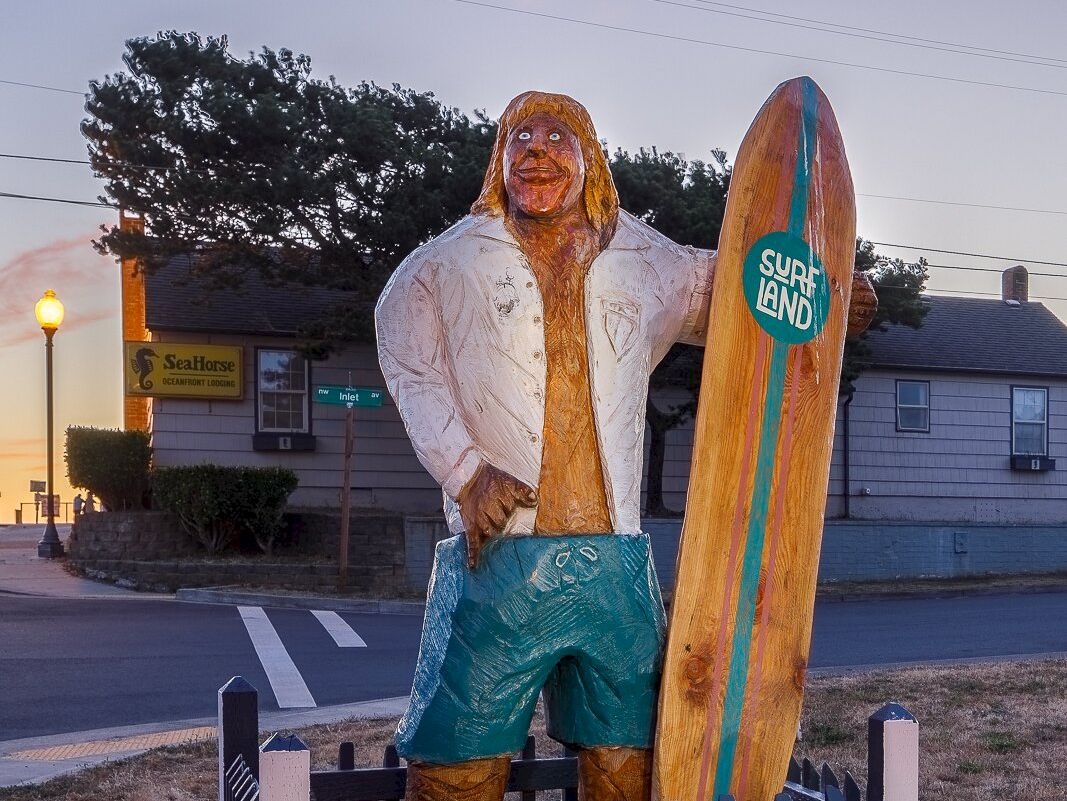 A wooden surfer statue holding a &ldquo;Sure Land&rdquo; surfboard stands in a yard behind a picket fence near a motel sign.