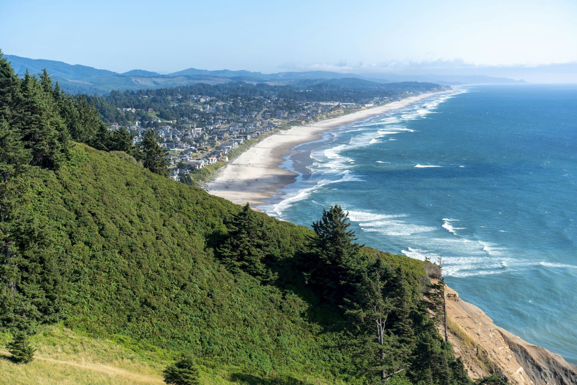 Scenic view of a forested hillside overlooking a coastal town and long sandy beach with waves rolling in.