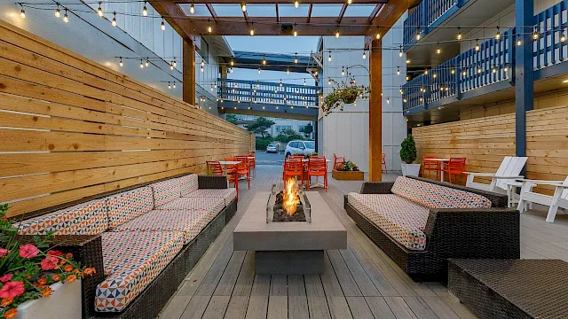 A cozy outdoor patio with string lights, wooden walls, cushioned benches, and a central fire pit.