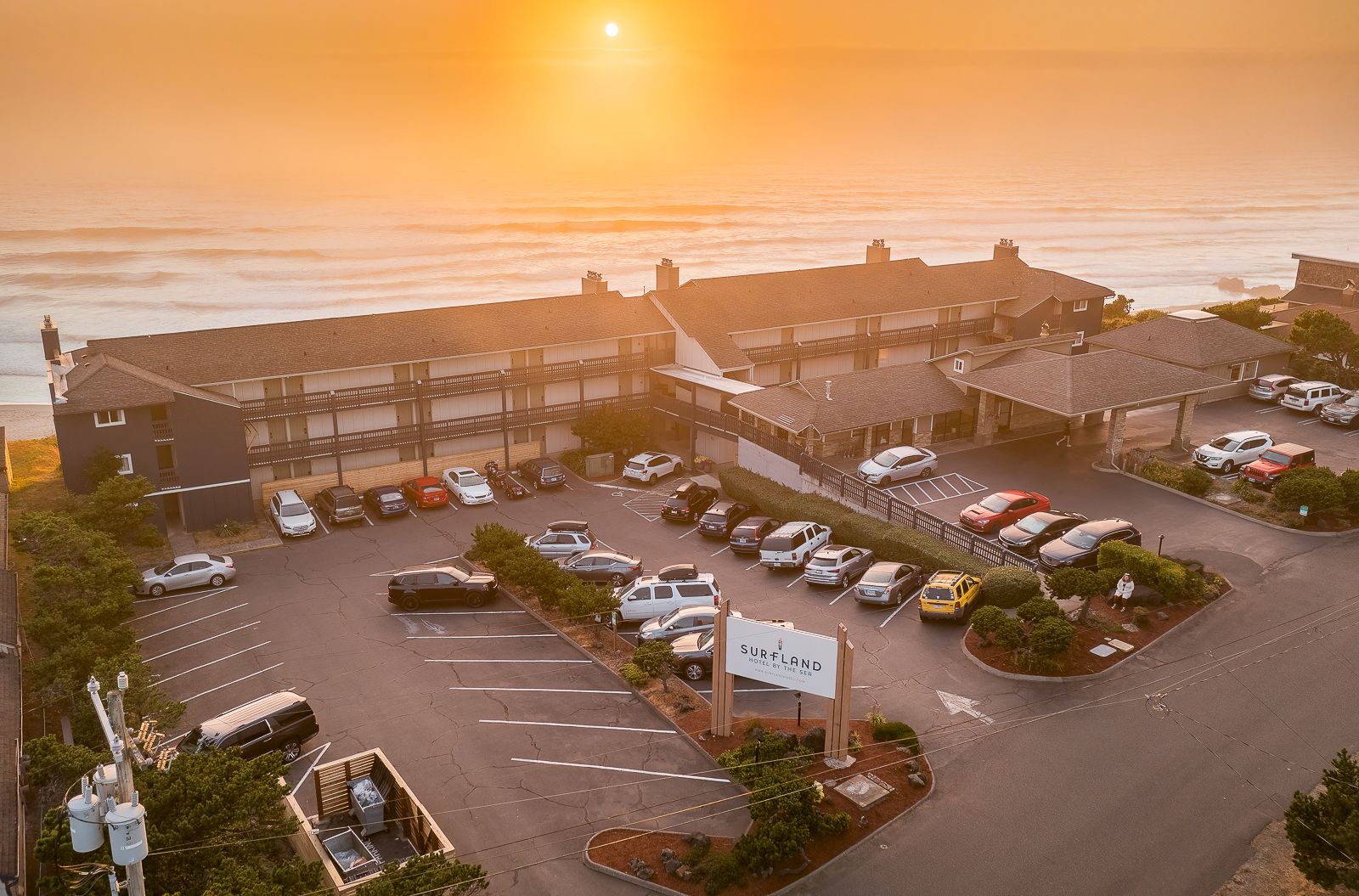 Aerial view of a beachside hotel, packed parking lot with cars, and the sunset over the ocean in the background.