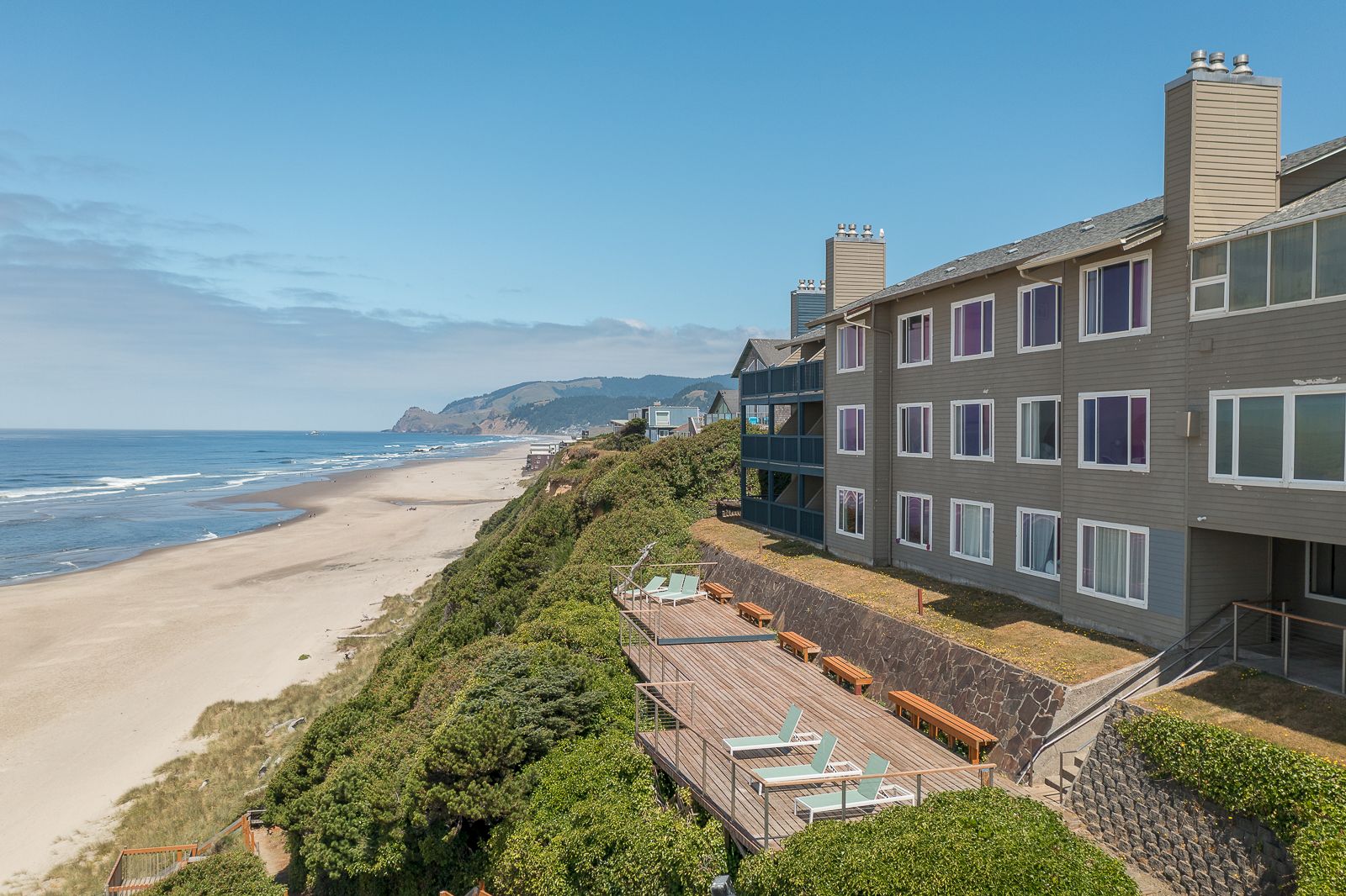 Oceanfront resort buildings with purple-trimmed windows overlook a sandy beach, waves, and a clear blue sky.