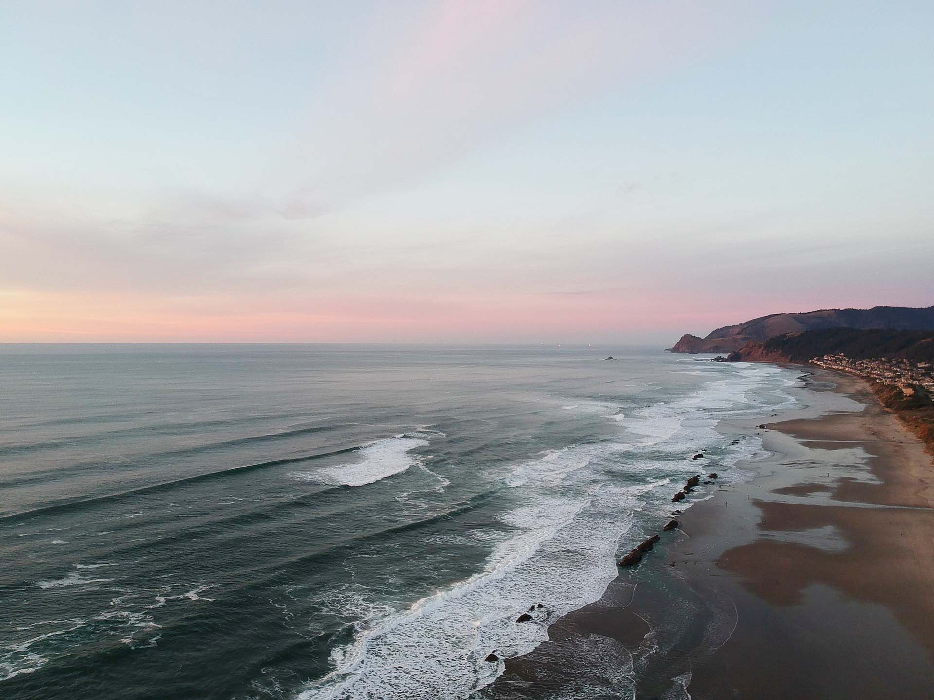 A quiet coastline at sunset with waves rolling onto a sandy beach and a cliff-lined shore under pastel clouds.